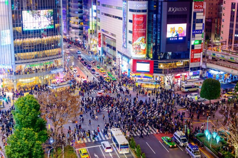 An aerial view of Shibuya Crossing in Tokyo at night, with numerous pedestrians crossing the street and illuminated buildings and advertisements surrounding the area. The vibrant scene is easily accessible for those with a Japan Rail Pass, making it a must-visit for travelers.