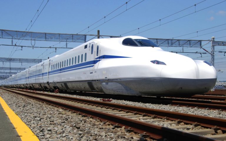 A white and blue high-speed train, eligible for travel with a Japan Rail Pass, glides on a track under overhead power lines, set against a clear blue sky.
