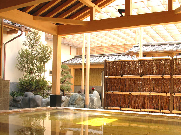A serene, traditional Japanese hot spring bath area with wooden structures, bamboo fencing, stones, and a roof partially allowing natural sunlight to filter through is easily accessible for travelers with a Japan Rail Pass.