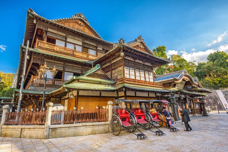 A historic multi-level wooden building with intricate architectural details stands majestically as rickshaws line up outside. People are conversing near the entrance, perhaps discussing their next destination on their JR Pass. The sky is clear and blue, perfectly framing the scene.