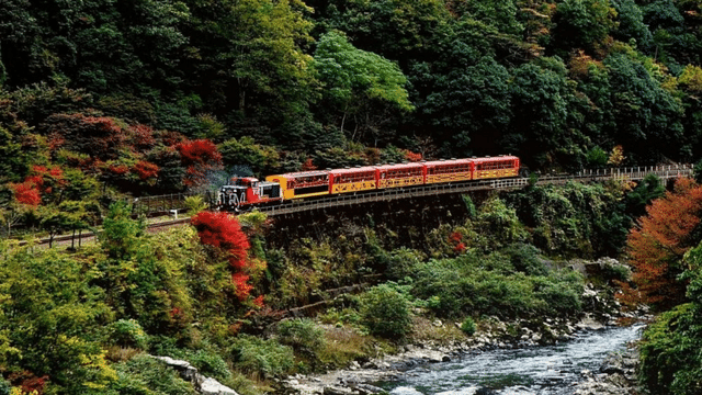 A red train travels over a bridge through a lush forest with green and red foliage, alongside a flowing river on a serene white day.