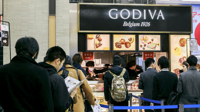 People queue in front of a Godiva store with a "Belgium 1926" sign. The store displays various chocolate products in glass cases and overhead images, perfect for celebrating White Day.