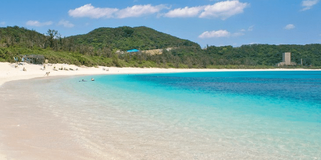 A tranquil beach with clear blue water, white sand, a few people relaxing on this serene white day, and green hills in the background under a partly cloudy sky.