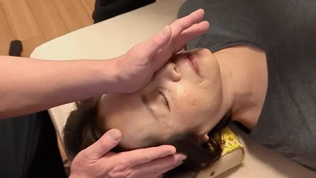 Person lying down with eyes closed receives a gentle Small Face Correction facial massage from another person's hands, head supported by a small cushion.