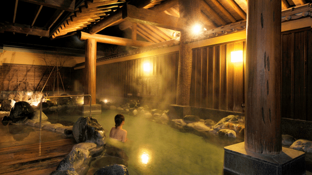 A person is sitting in an outdoor hot spring bath at a traditional Japanese onsen during the evening. The surrounding area features wooden structures and warm lighting.
