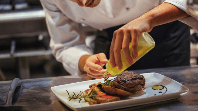 A Michelin-rated chef in a white coat carefully drizzles oil over a plated dish of steak and garnishes in a kitchen setting.