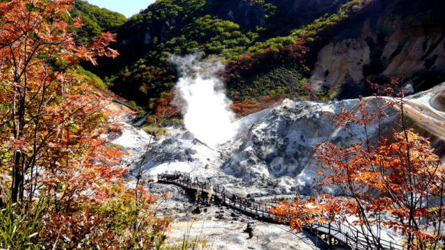 A natural hot spring emits steam amidst autumn foliage in a mountainous landscape, with a boardwalk leading up to it—truly capturing the essence of onsens in Japan.