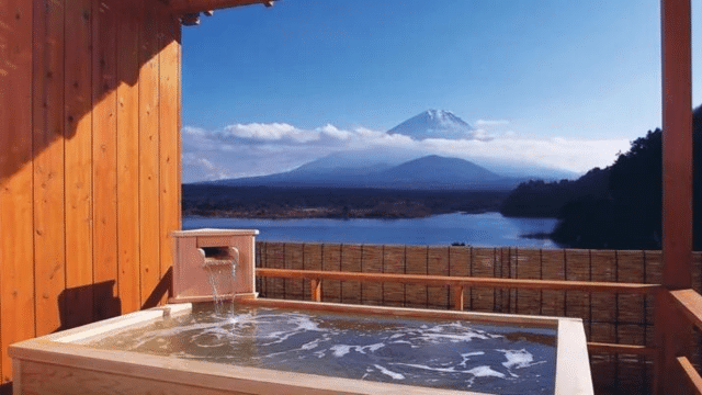 Outdoor hot spring bath with wooden framing overlooking a calm lake and distant snow-capped mountain under a clear blue sky—experience the tranquility of traditional onsens in Japan.