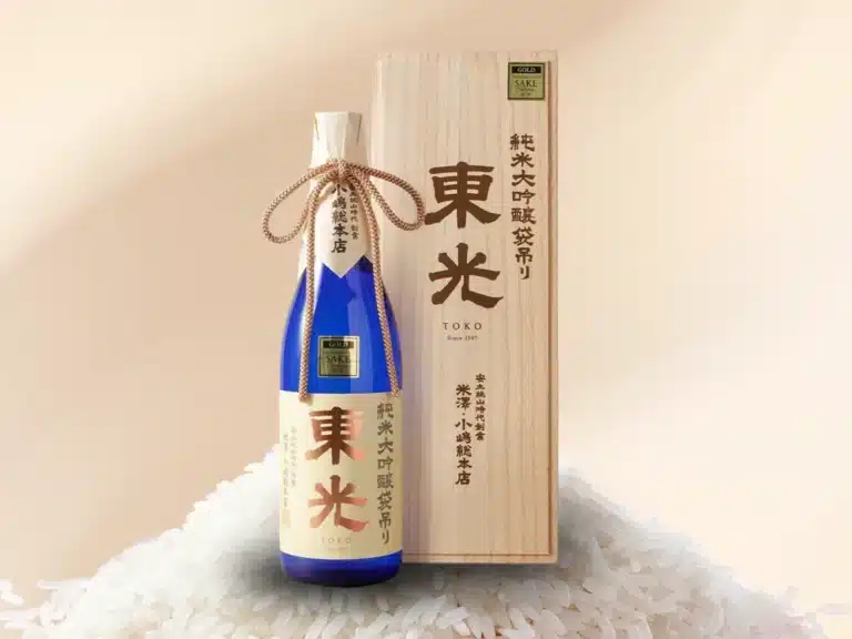 A blue bottle of TOKO sake with Japanese writing stands next to a wooden gift box, both displayed on a bed of rice grains—an elegant pairing perfect for a couples’ spa package experience.