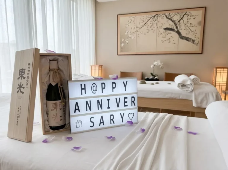 A lightbox sign reading "Happy Anniversary" sits on a bed with an anniversary gift—a sake bottle in a wooden box—surrounded by petals and rolled towels in a modern, softly lit room.
