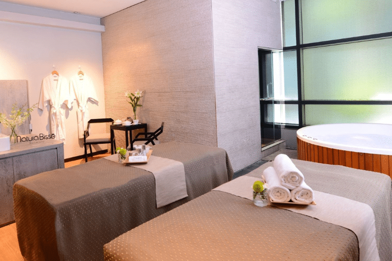 A spa room in Singapore with two massage tables, fresh towels, a small bouquet on each table, a corner jacuzzi, and robes hanging on the wall next to a small table and chair. Perfect for those seeking massages in Singapore.