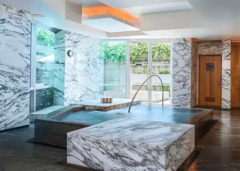 A luxurious indoor spa area featuring a small pool with a metal handrail, surrounded by marble walls and floors, adjacent to a wooden sauna door, with large windows overlooking greenery—perfect for enjoying massages in Singapore.