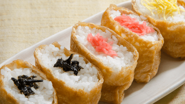A plate with pieces of inari sushi, featuring rice in fried tofu pouches and toppings such as shredded nori, pickled ginger, and yellow shredded egg