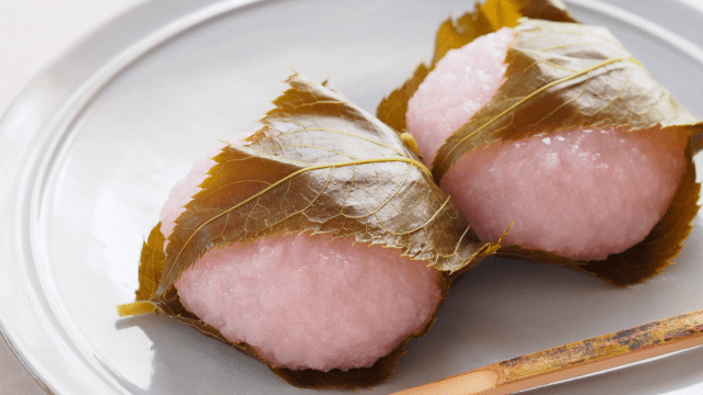 Two pink rice cakes wrapped in cherry leaves are placed on a white plate with a wooden utensil beside them.