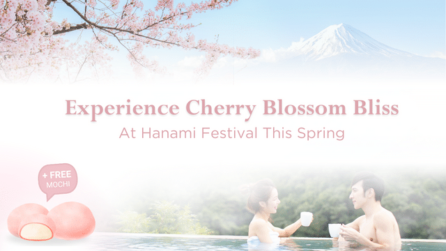 Two people in a hot spring enjoy a cherry blossom view with Mount Fuji in the background.