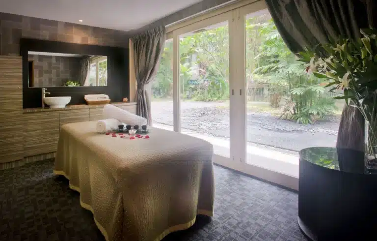 A spa treatment room with a massage table, rolled towels, flower petals, and a tea set, next to large glass doors overlooking a garden—perfect for relaxing massage Singapore experiences.