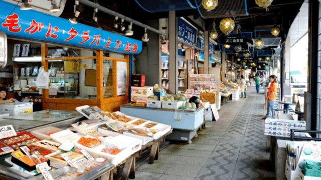 A bustling Japan market with various seafood stalls, boxes of produce, and people shopping. Japanese signage is visible above the stalls.