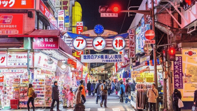 A bustling Japan night market with brightly lit signs and numerous stalls, crowded with people shopping and walking.