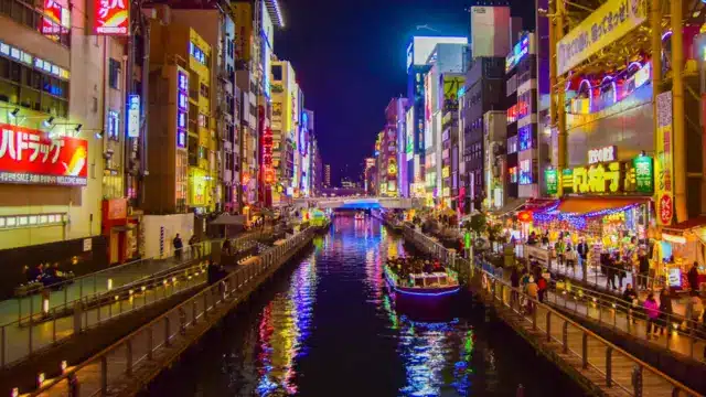 A vibrant city canal at night with illuminated buildings, neon signs, and boats on the water, surrounded by busy walkways and crowds reminiscent of a Japan night market.