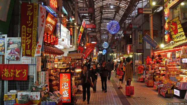 Busy indoor Japan night market with various colorful stalls showcasing food items, snacks, and goods. Shoppers walk through the narrow aisle under a ceiling adorned with blue decorative lights. Signs are written in Japanese.