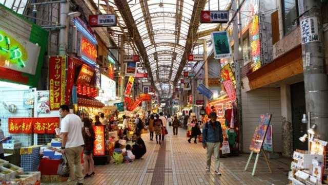 A bustling indoor market reminiscent of a Japan night market, with people walking and shopping and vibrant storefronts displaying various goods and signs in a covered arcade.