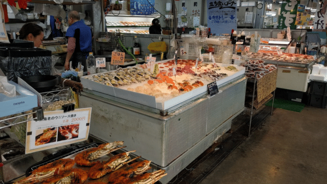 A bustling seafood market stall with a variety of fish and shellfish on ice evokes the lively atmosphere of a Japan market. In the background, people handle products and make transactions, while various signs with prices are visible.