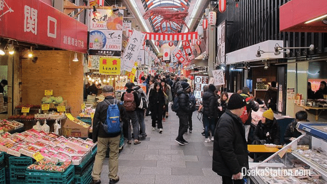 A bustling indoor market in Osaka features stalls with food, produce, and other goods. Shoppers and vendors are present, and various signs are displayed throughout.