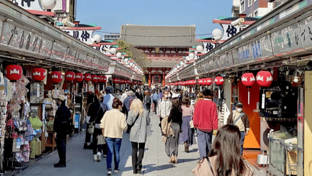 A crowd of people walks through a market, lined with shops and Japanese lanterns. A traditional building with a large roof is visible in the background.