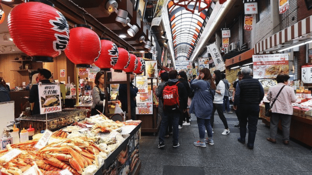 People stroll through a bustling indoor market, with various stalls selling seafood and other goods. Red lanterns adorn the ceiling, and signs are displayed throughout the vibrant space.