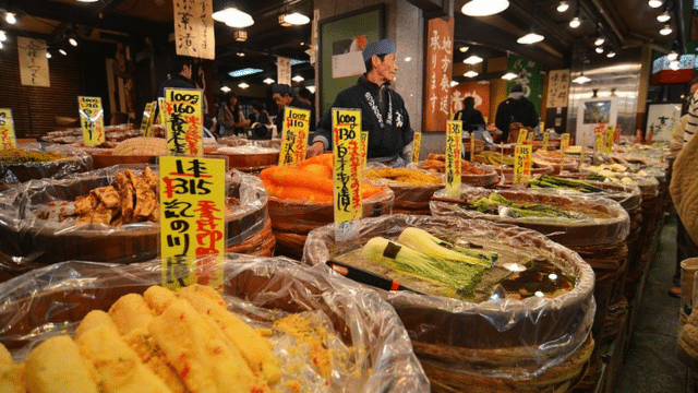A bustling Japan night market scene showcases a variety of pickled vegetables in large barrels labeled with prices and descriptions in Japanese. A vendor and several eager customers are visible in the background, adding to the vibrant atmosphere.