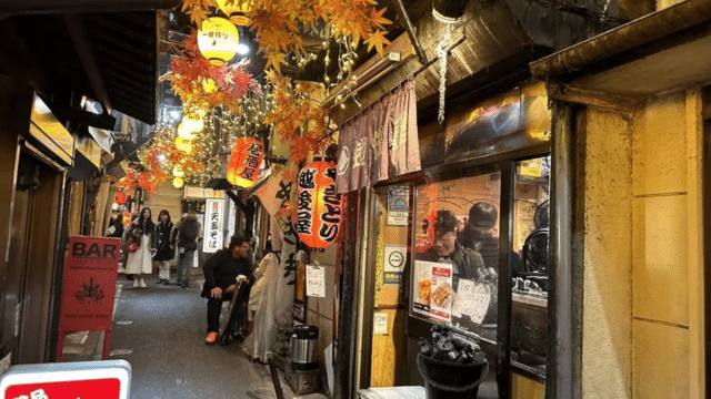 A narrow alley lined with small restaurants and vibrant lanterns, with people walking and dining, captures the essence of a Japan night market. The scene is decorated with artificial autumn leaves, creating a warm and inviting atmosphere.
