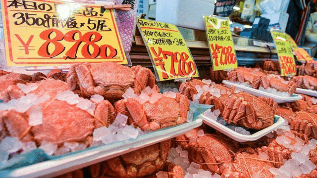Trays of red crabs on ice are displayed in a Japan night market. Yellow and red price signs indicate the cost of the crabs in Japanese yen.