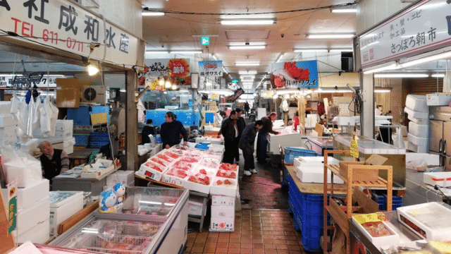 An indoor seafood market reminiscent of a bustling Japan night market, with various vendors selling fish and other seafood products. Customers are browsing and interacting with sellers.
