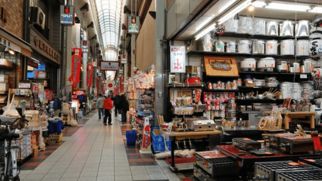 Indoor market with various shops selling kitchenware, food items, and other goods. Shoppers walk along the central aisle under a high, curved ceiling.