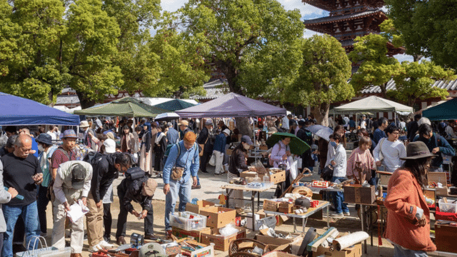 A bustling outdoor Japan night market with people browsing various stalls under canopies, surrounded by greenery and a multi-story pagoda glowing in the background.