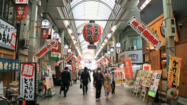 A bustling Japan market with people walking through, lined with shops displaying various colorful signs and banners under a glass-paneled ceiling.