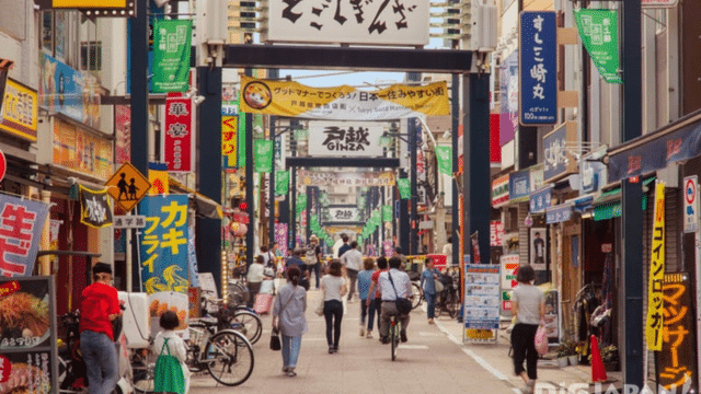 A bustling Japanese shopping street, echoing the vibrant spirit of a Japan market, with colorful signs and banners overhead. People walk, bicycle, and shop along the lively lanes lined with various shops and eateries.