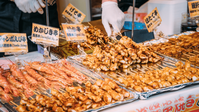 A vendor wearing gloves handles skewers of grilled seafood and meat at a Japan food stall, with various skewers including shrimp, squid, and chicken displayed on trays with signs.