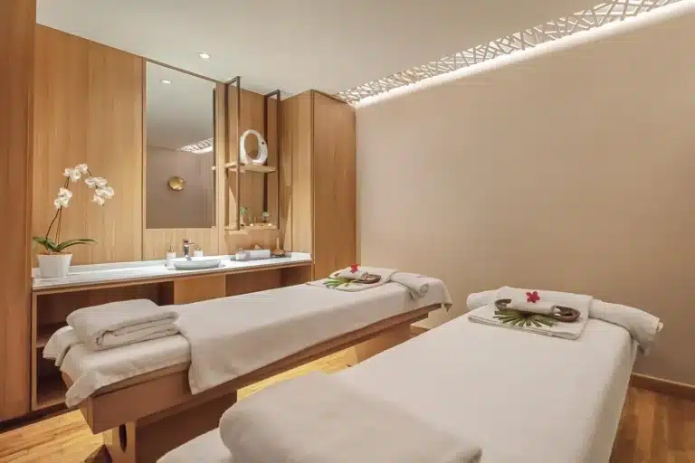 Experience tranquility in this modern spa room, perfect for massage Singapore seekers, featuring two massage tables with crisp white linens, decorative flowers, a potted orchid, and a stylish sink area with wooden accents.