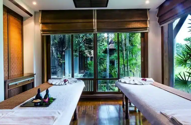 Two massage tables with white towels and flowers are set up in a serene spa room with large windows overlooking a lush garden, offering a tranquil massage Singapore experience.