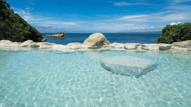 Clear swimming pool with submerged rock in the center, surrounded by a stone wall. In the background, a view of the ocean and lush greenery under a blue sky with scattered clouds evokes the tranquil beauty of onsens in Japan.