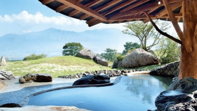 Outdoor hot spring with clear water, surrounded by rocks, trees, and green grass, under a wooden roof. In the background, a mountain range can be seen against a partly cloudy sky—a serene scene reminiscent of an onsen in Japan.