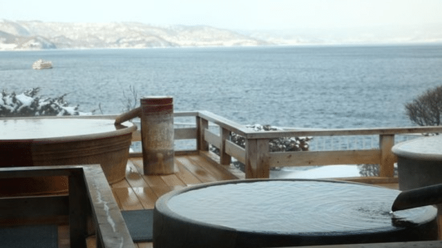Experience the serenity of outdoor onsen in Japan, featuring hot spring baths that overlook a calm body of water with a distant boat on the horizon. Snow-covered mountains provide a stunning backdrop, enhancing this tranquil setting.