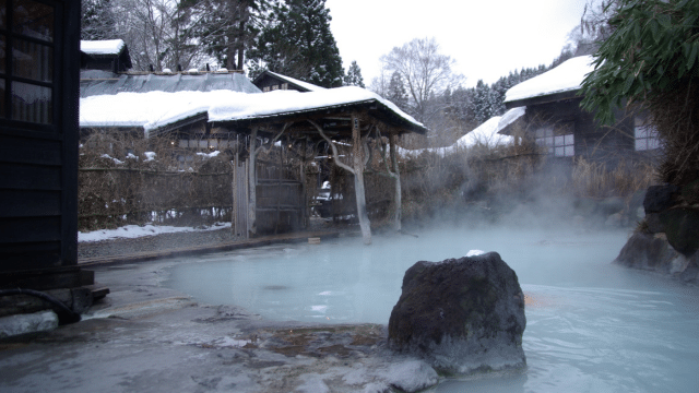 Outdoor onsen in Japan with steam rising, surrounded by snow and traditional wooden buildings.