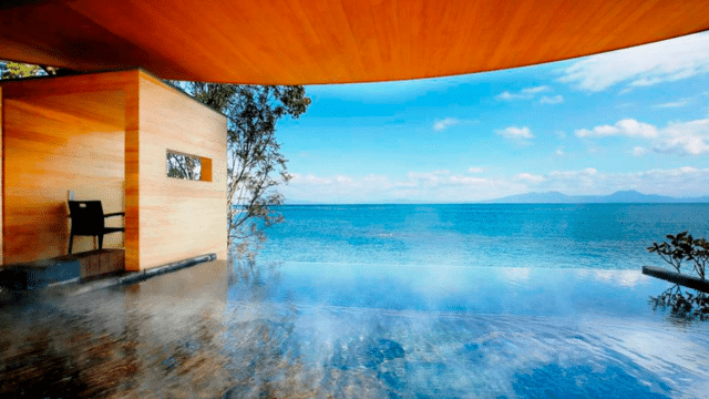 A modern wooden structure adjacent to a tranquil infinity pool overlooks a vast body of water with mountains in the distance under a clear blue sky, reminiscent of serene onsens in Japan.