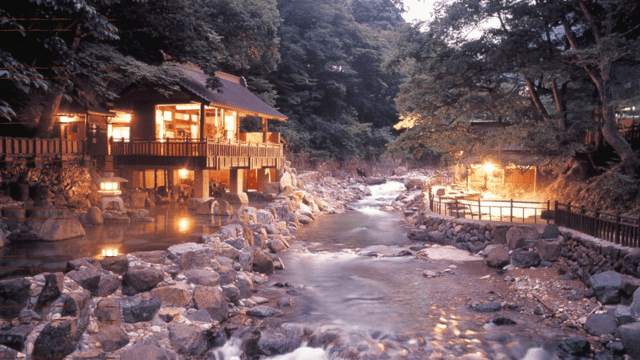A serene riverside hot spring resort at dusk features a wooden building with warm lighting, surrounded by trees and rocky terrain, next to a gently flowing stream, reminiscent of the tranquil onsens in Japan.