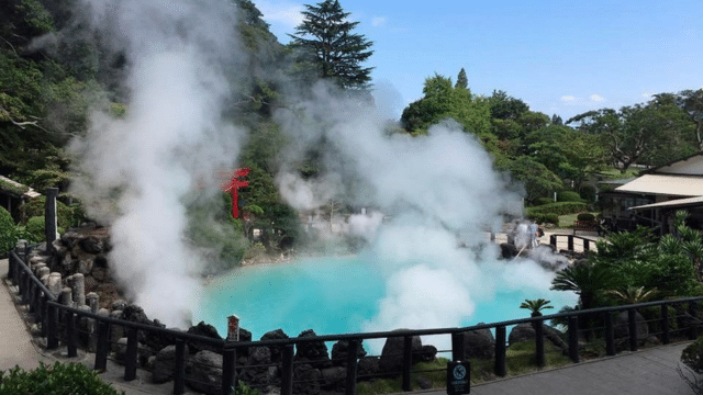 Geothermal hot spring with steam rising, surrounded by lush greenery and a fence. A red torii gate is partially visible in the background, capturing the serene beauty typical of onsens in Japan.