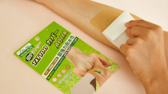 A person applies a skin-colored sticker over a tattoo on their forearm. Packaging labeled "Tattoo Cover Aqua" is visible next to the arm.