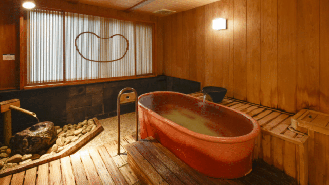 A traditional Japanese bathhouse room with a red bathtub, wooden floors, stone accents, and wooden paneled walls awaits you.