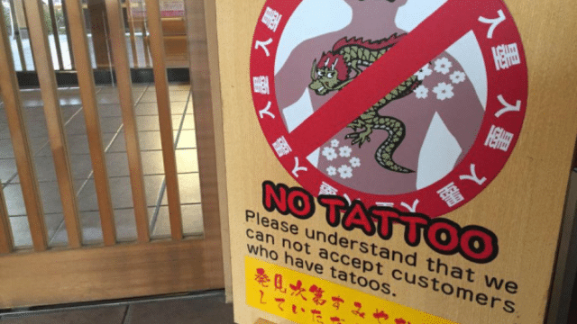 Sign on a door with a red circle and a line through a tattooed figure, stating, "No Tattoo. Please understand that we cannot accept customers who have tattoos at this tattoo friendly onsen.